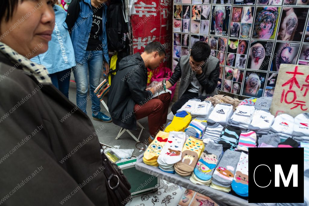 duas pessoas sentadas numa rua, sendo um tatuador e a pessoa que está sendo tatuada, outras observando ao redor. 100 CNY é parte da série outras estórias por Ca Mila