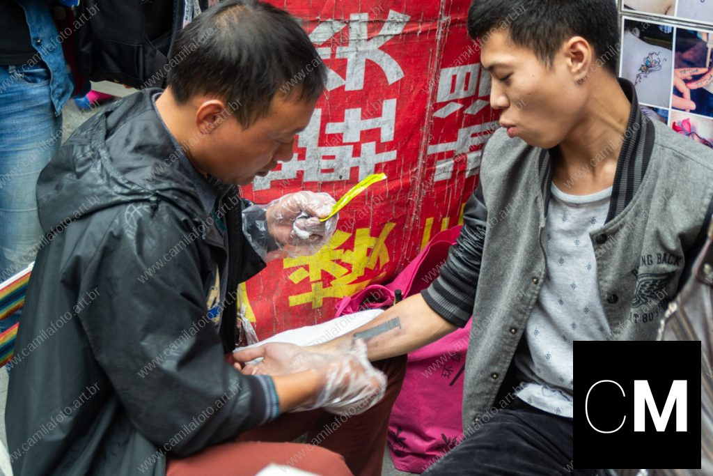 duas pessoas sentadas numa rua, sendo um tatuador e a pessoa que está sendo tatuada. 100 CNY é parte da série outras estórias por Ca Mila