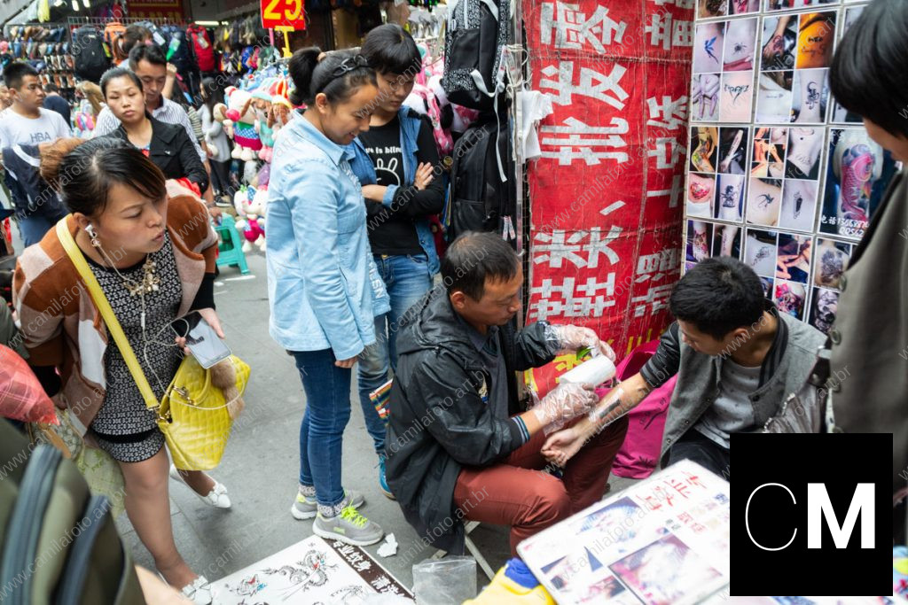duas pessoas sentadas numa rua, sendo um tatuador e a pessoa que está sendo tatuada, outras observando ao redor. 100 CNY é parte da série outras estórias por Ca Mila