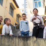 uma criança com roupas amarelas no canto esquerdo fazendo uma careta, uma criança com roupas brancas olhando para o lado, uma outra criança com as mãos no bolso olhando para a frente e uma mãe com o filho apontando para a câmera. 1,2,3 e... é parte da série emoções por Ca Mila